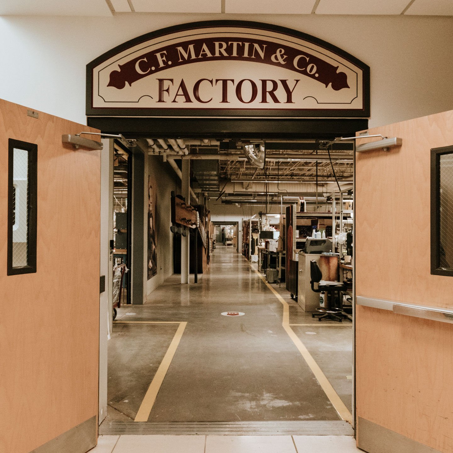 Looking at two doors opening into a factory with concrete floors and yellow safety paint lining the walkway. The sign above the door says "C.F.. Martin & Co. Factory"