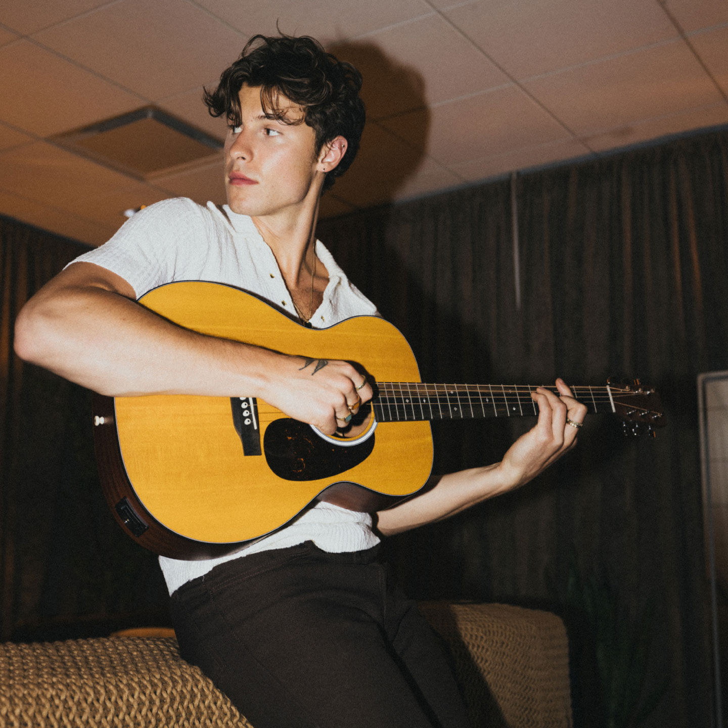 Shawn Mendez playing a 000 Martin in a white shirt while leaning against a mustard yellow couch