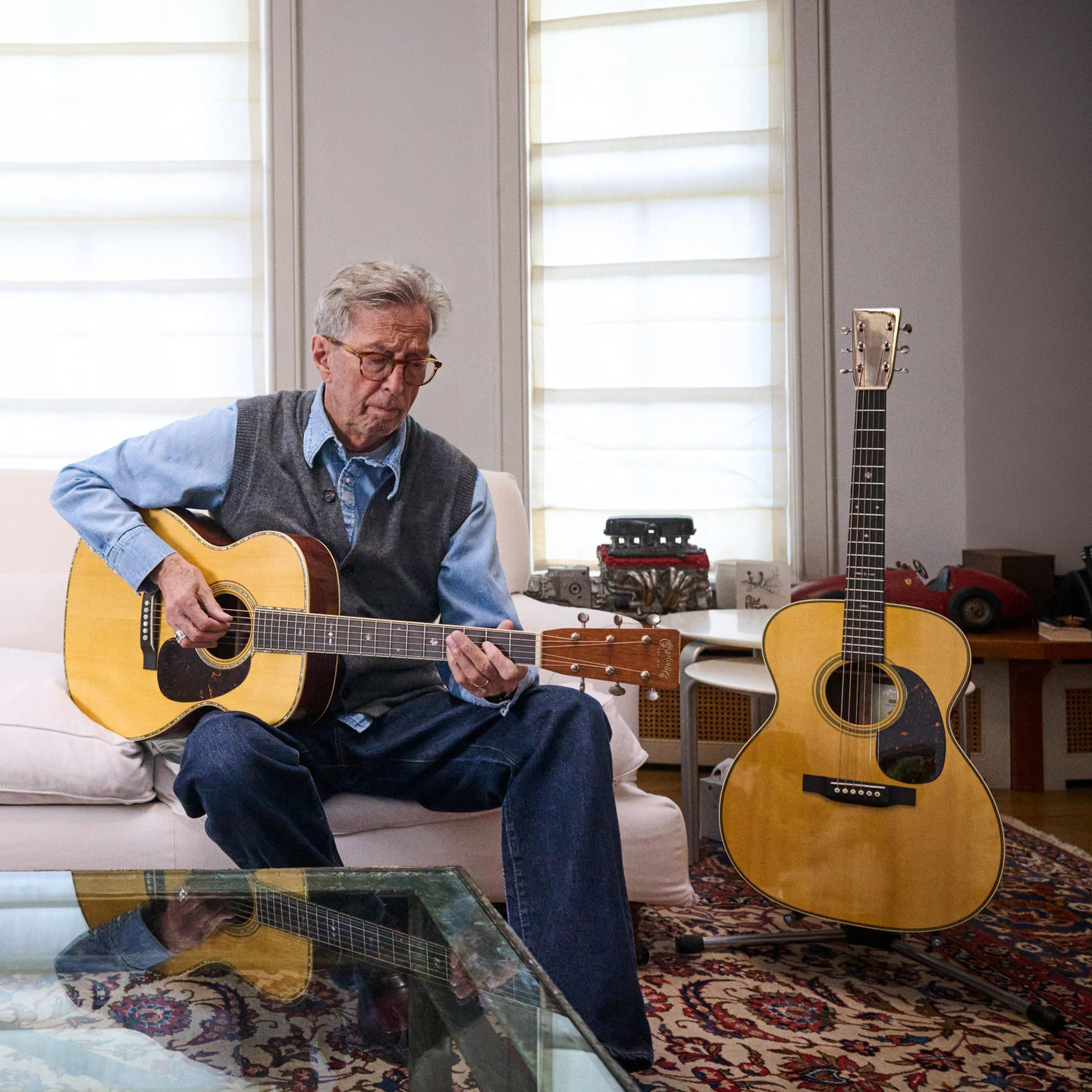 Eric Clapton sitting on a white couch playing one of his model guitars, while the other sits on a stand behind him