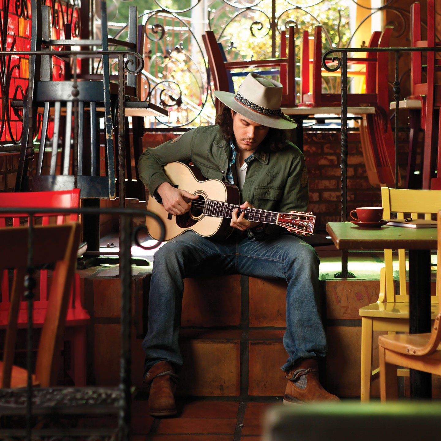 John Mayer wearing a brimmed hat and playing a Martin acoustic while sitting in a restaurant. Chairs are stacked on top of the table.