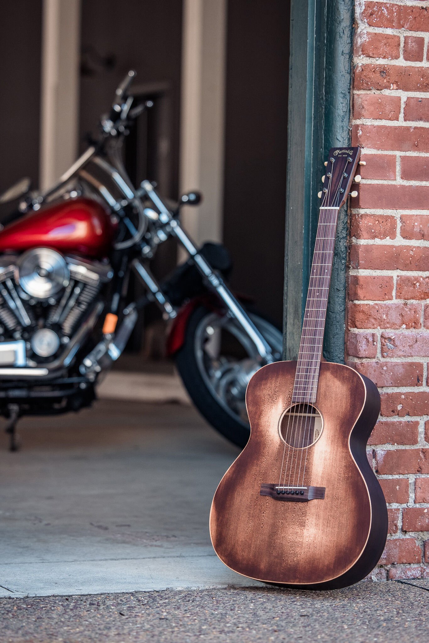 Photo of an acoustic guitar resting against a brick wall with a red motorcycle in the background