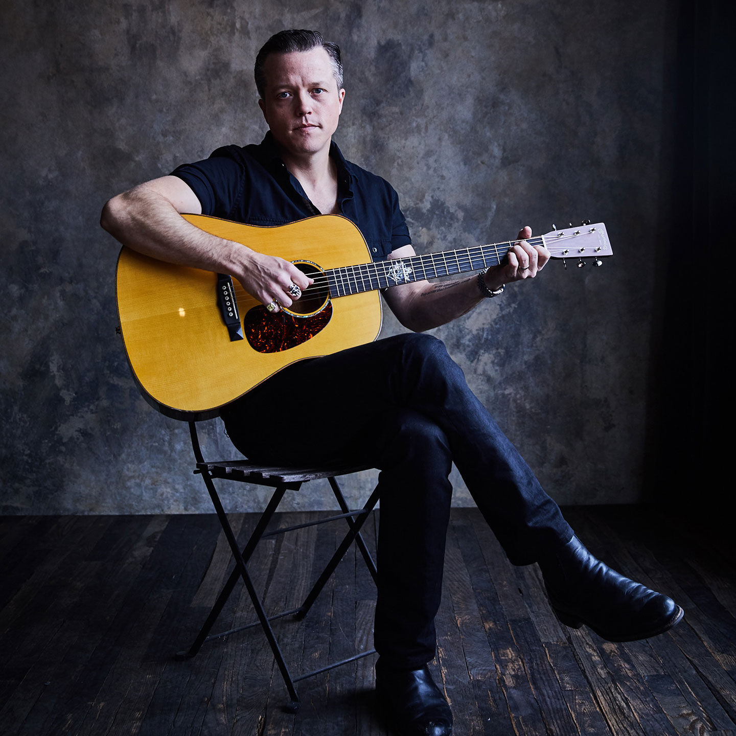 Jason Isbell wearing dark colors holding a Martin Dreadnought guitar while sitting in a folding chair