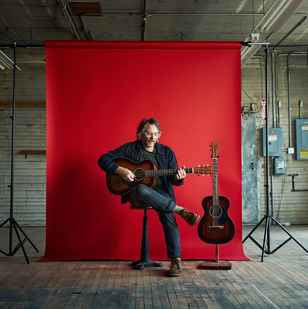 Jeff Tweedy in front of a red backdrop with his acoustic guitars Jeff Tweedy in front of a red backdrop with his acoustic guitars
