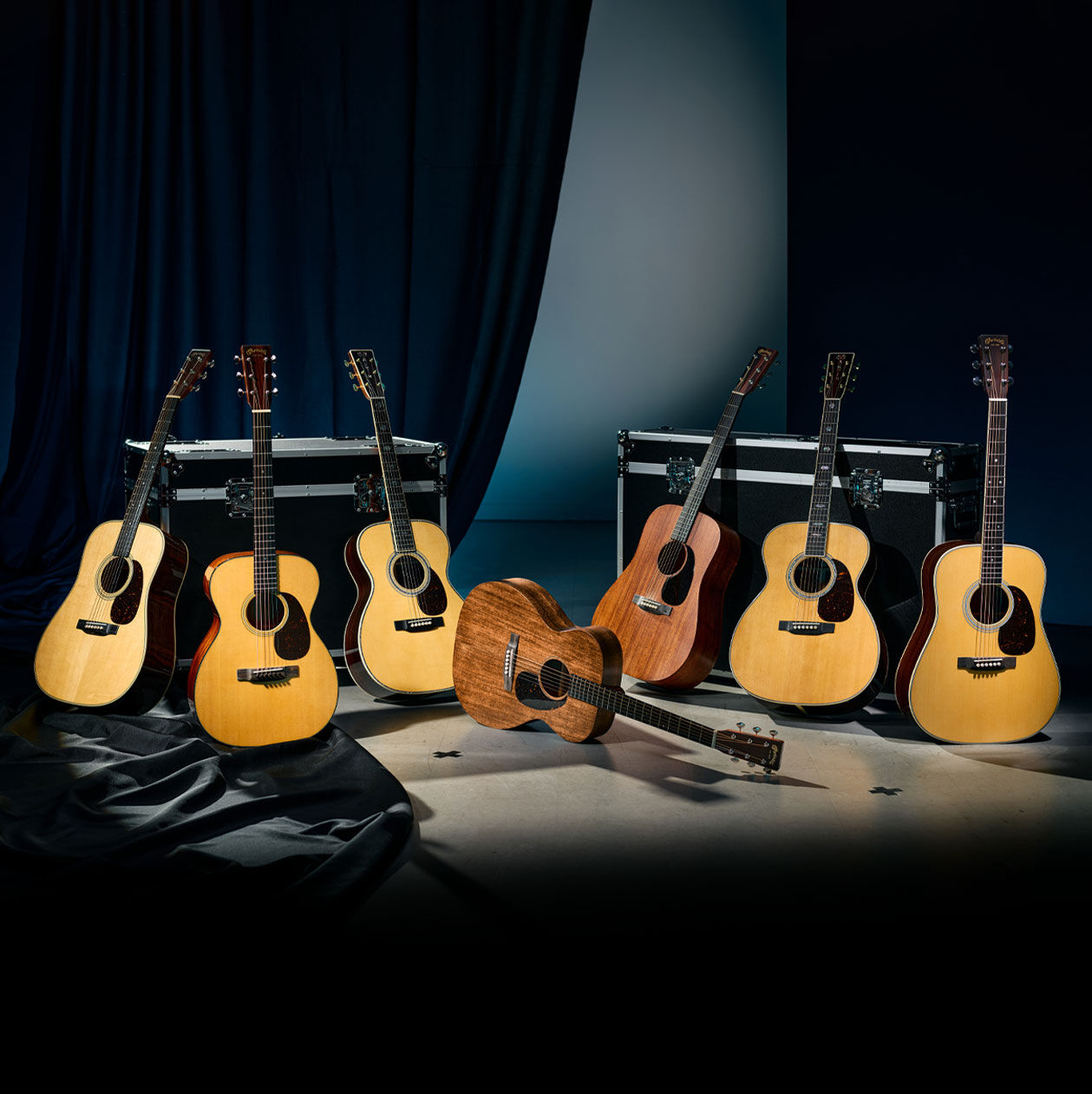 A group of 7 different acoustic guitars in a black room.